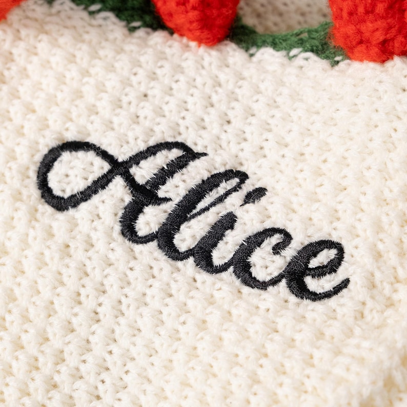 Puede incluir: Un tejido de punto color crema con el nombre Alice bordado en letra cursiva negra. El borde superior del textil est&aacute; decorado con una hilera de peque&ntilde;as esferas rojas texturizadas unidas a tallos y hojas de ganchillo de color verde.
