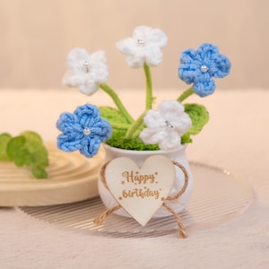 May include: A white ceramic pot with a heart-shaped wooden sign that says "Happy birthday" is filled with a bouquet of crocheted flowers. The flowers are white and blue.