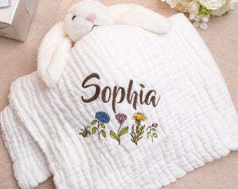 Op de afbeelding: Witte katoenen babydeken met een wit konijn eraan bevestigd. De deken heeft een geborduurde naam "Sophia" in bruin en een rij van kleurrijke geborduurde bloemen.