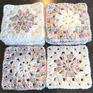 May include: Four square crochet motifs arranged in a grid. Two motifs feature a white center with a pastel variegated border, while the other two feature a pastel variegated center with a white border. The yarn colors include white, pink, blue, and yellow.