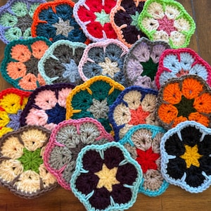 May include: A collection of colorful, handmade crochet coasters in various shades, including red, orange, yellow, green, blue, purple, and pink. Each coaster features a floral design with a central starburst pattern. The coasters are arranged on a wooden surface.