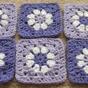 May include: Six crocheted squares in shades of purple and white. Each square features a white flower design in the center.