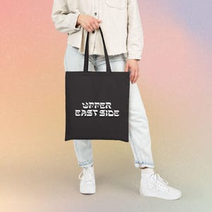 May include: A black tote bag featuring the white text "UPPER EAST SIDE" printed in a bold, stylized font on the front. The bag has two black shoulder straps and is constructed from a solid, dark fabric material with a rectangular shape.