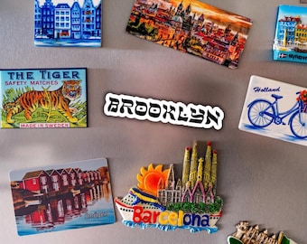 May include: A collection of rectangular and shaped souvenir magnets featuring various international cityscapes and landmarks, including Malmö, Amsterdam, Prague, Saint Petersburg, The Tiger, Brooklyn, Holland, Smögen, Barcelona, Croatia, and Venezia.