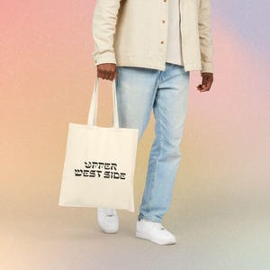 May include: A cream-colored canvas tote bag featuring the black text "UPPER WEST SIDE" printed on the front. The bag has two long shoulder straps and a rectangular body, shown held against a light-colored shirt and blue denim jeans.