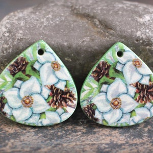 May include: Two teardrop-shaped pendants with a floral design. Each pendant features white flowers with yellow centers, brown pinecones, and green leaves against a white background. The edges are green, and each has a hole at the top.