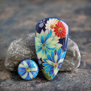 May include: Two blue and white floral patterned polymer clay cabochons. The larger cabochon is shaped like a teardrop and the smaller cabochon is round. Both cabochons have a blue, white, and gold floral pattern.