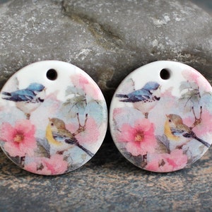 May include: Two round ceramic pendants with a white background and a floral design. The design features pink flowers, green leaves, and three birds. One bird is blue and white, and the other two are yellow with brown wings.