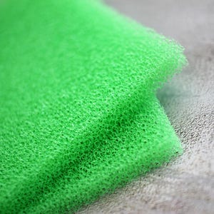 May include: A bright green, rectangular foam filter pad featuring a highly porous, open-cell structure. The material is lightweight and flexible, displaying a consistent, uniform texture throughout its surface area with visible interconnected air pockets.
