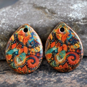 May include: Two teardrop-shaped ceramic pendants with a colorful paisley design. The pendants are orange, blue, green, and gold with a black background. Each pendant has a hole at the top for stringing.