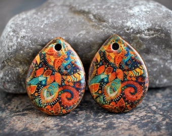 Puede incluir: Dos colgantes de cerámica en forma de lágrima con un diseño paisley colorido. Los colgantes son de color naranja, azul, verde y dorado sobre un fondo negro. Cada colgante tiene un agujero en la parte superior para encordar.