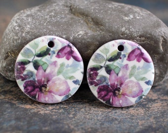Puede incluir: Dos colgantes redondos de cerámica con un diseño floral. Los colgantes son blancos con una impresión de estilo acuarela de flores rosas y moradas y hojas verdes. Cada colgante tiene un pequeño agujero en la parte superior para colgar.