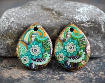 Puede incluir: Dos colgantes en forma de lágrima con un diseño floral. Los colgantes presentan un patrón floral turquesa, verde y morado sobre un fondo crema. Los bordes son de color marrón cálido y cada uno tiene un pequeño agujero en la parte superior para colgar.