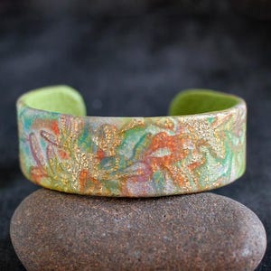 Könnte beinhalten: Ein Armband aus Polymer-Ton mit grünem, goldenem und orangefarbenem Muster. Das Armband ist aus einem glatten, glänzenden Material gefertigt und hat ein einzigartiges, abstraktes Design.