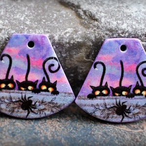 May include: Two purple and pink painted ceramic pendants with a black cat design. Each pendant features three black cats with yellow eyes peeking over a fence with a spider in the foreground.