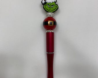 Può includere: Una penna rossa con un personaggio verde del Grinch che indossa un cappello da Babbo Natale in cima. La penna ha una palla rossa e dorata con strass intorno.