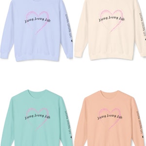 May include: Four crewneck sweatshirts in different colors: light blue, beige, light green, and peach. Each sweatshirt features a pink heart with a sparkly texture and the text "Living Loving Life" in black lettering. The text is repeated on the sleeves of each sweatshirt.