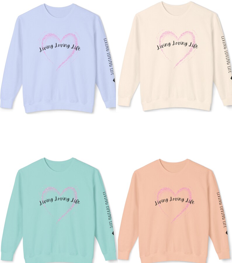 May include: Four crewneck sweatshirts in different colors: light blue, beige, light green, and peach. Each sweatshirt features a pink heart with a sparkly texture and the text "Living Loving Life" in black lettering. The text is repeated on the sleeves of each sweatshirt.