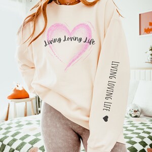 May include: A cream-colored sweatshirt with a pink glitter heart and the text "Living Loving Life" on the front. The same text is repeated on the sleeve with a small heart.