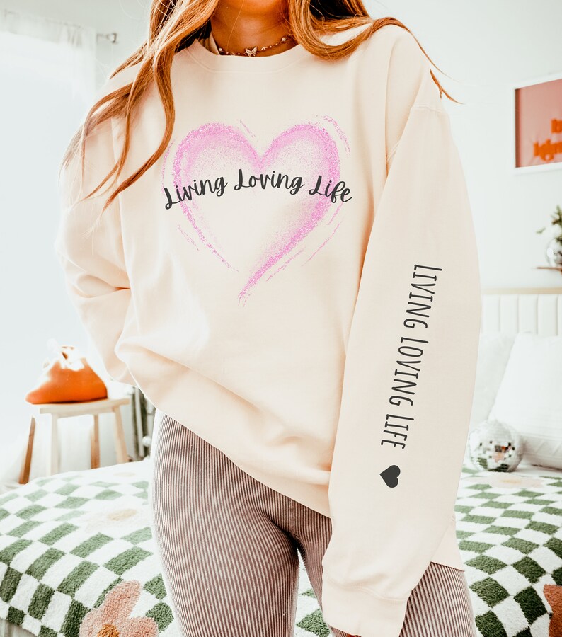 May include: A cream-colored sweatshirt with a pink glitter heart and the text "Living Loving Life" on the front. The same text is repeated on the sleeve with a small heart.