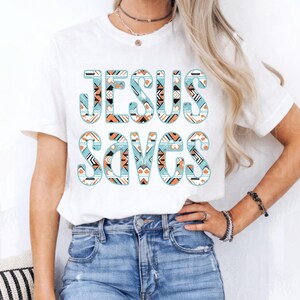 May include: White t-shirt with the words "JESUS SAVES" in large, stylized letters. The letters are filled with a geometric pattern in shades of blue, orange, and white. The shirt is a casual, everyday wear item.