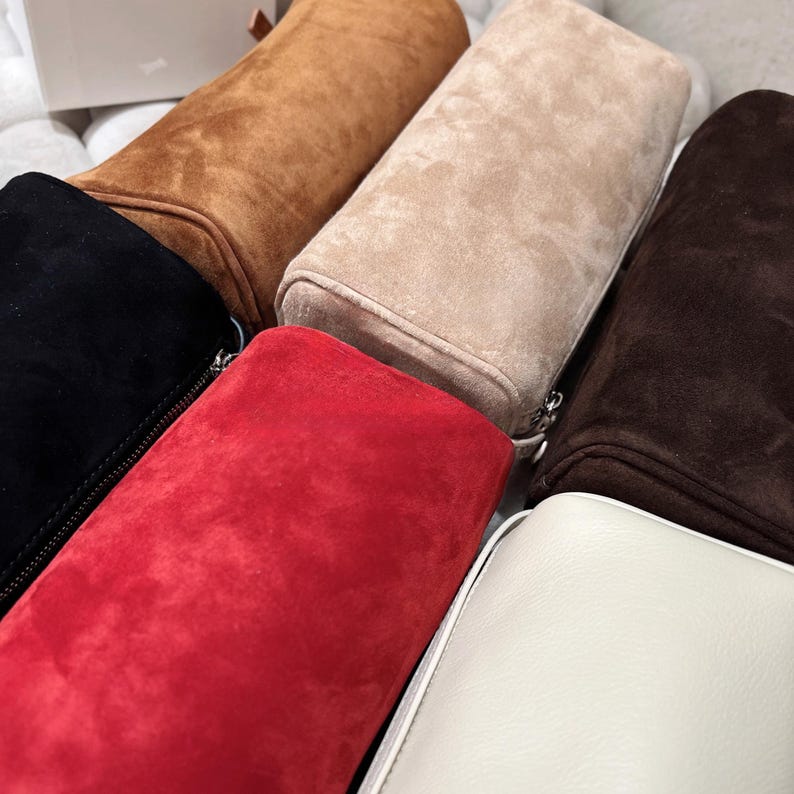 May include: An assortment of suede bags in various colors, including black, brown, tan, red, and white. Each bag has a rectangular shape with a rounded top and a zipper closure. The bags are arranged closely together.