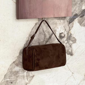 May include: A brown suede shoulder bag with a rectangular shape and a silver zipper. The bag has a thin, adjustable strap and is hanging against a marble-patterned wall.