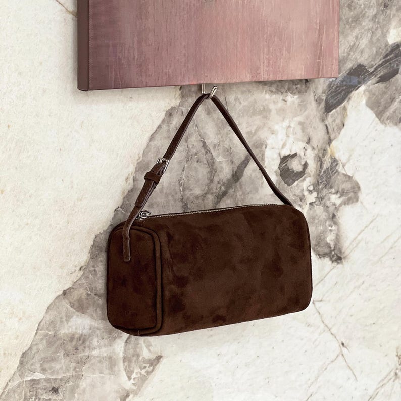 May include: A brown suede shoulder bag with a rectangular shape and a silver zipper. The bag has a thin, adjustable strap and is hanging against a marble-patterned wall.