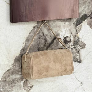 May include: A beige suede shoulder bag with a rectangular shape and a zipper closure. The bag has a thin strap and is hanging from a hook. The background features a marble-like wall and a dark brown rectangular object.