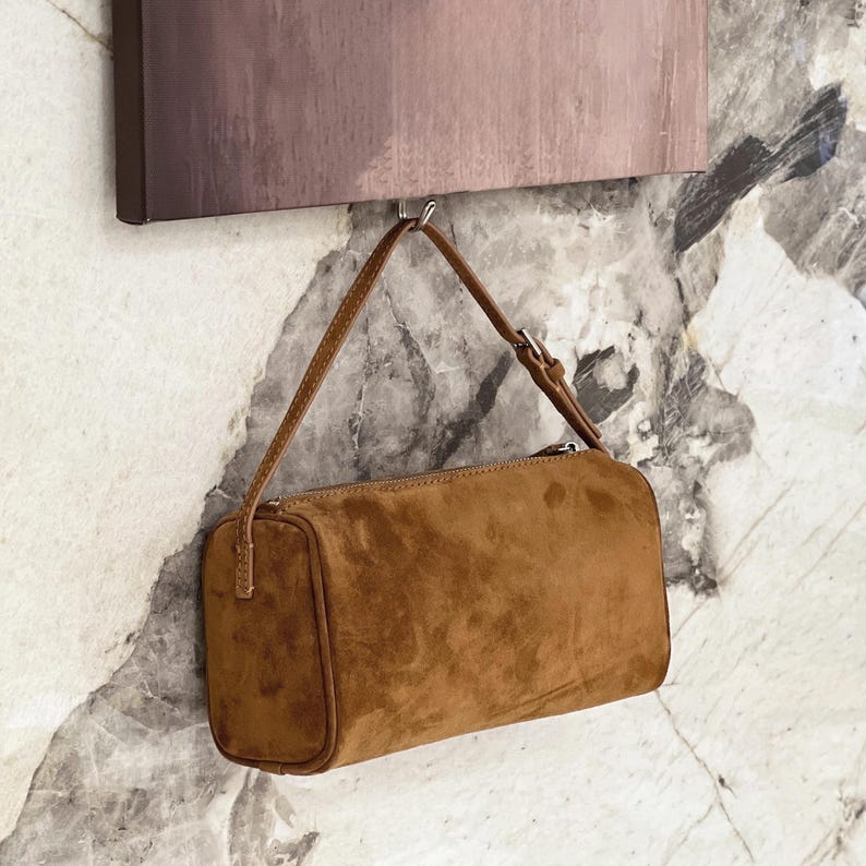 May include: A brown suede shoulder bag with a rectangular shape and a thin strap. The bag has a zipper closure and a small buckle on the strap. The bag is hanging from a hook.