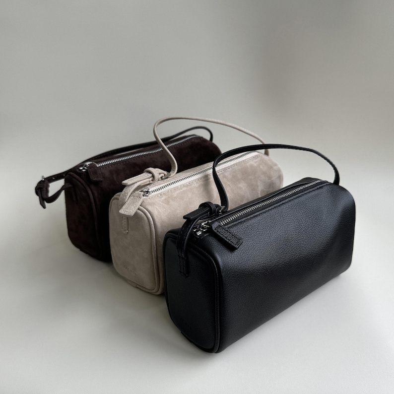 May include: Three small, rectangular handbags in a row. The bags are in shades of brown, beige, and black. Each bag has a zipper closure and a small handle. The black bag has a smooth leather texture, while the other two appear to be suede.