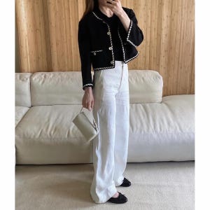 May include: A black jacket with white trim and gold buttons, paired with white wide-leg pants and black flats. A small, light-colored handbag is held in the hand. The outfit is worn in front of a white sofa and a wooden wall.