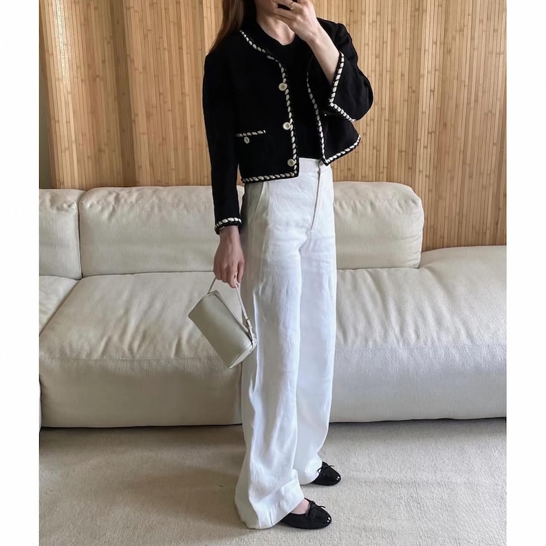 May include: A black jacket with white trim and gold buttons, paired with white wide-leg pants and black flats. A small, light-colored handbag is held in the hand. The outfit is worn in front of a white sofa and a wooden wall.
