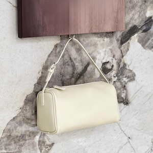 May include: Cream-colored rectangular handbag with a shoulder strap. The bag is hanging from a metal hook against a marble-patterned wall. The bag has a zipper closure and a small, rectangular design.