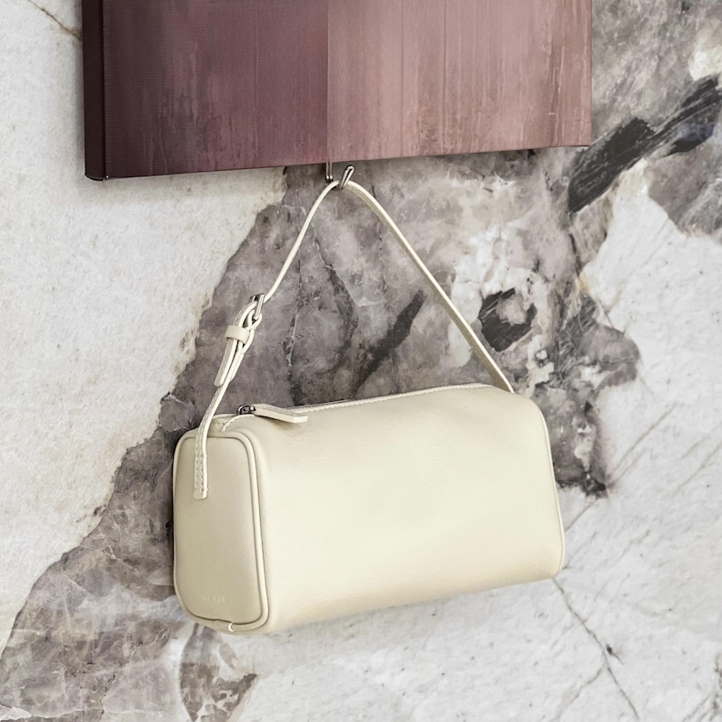 May include: Cream-colored rectangular handbag with a shoulder strap. The bag is hanging from a metal hook against a marble-patterned wall. The bag has a zipper closure and a small, rectangular design.