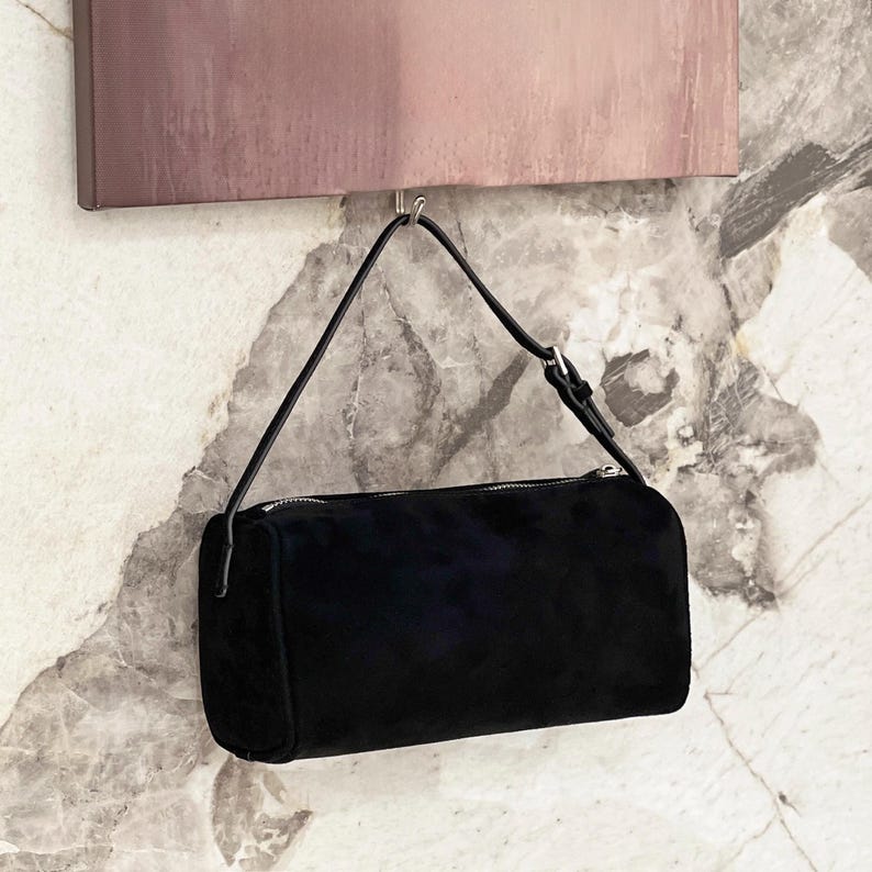 May include: A black suede shoulder bag with a rectangular shape and a zipper closure. The bag has a thin, black strap and is hanging from a metal hook. The background features a marble-patterned wall and a muted pink artwork.