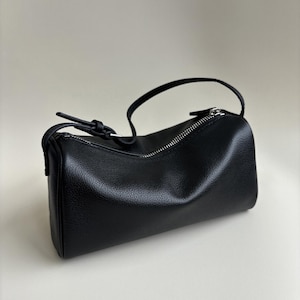 May include: A black, cylindrical handbag with a zipper closure and a small handle. The bag is made of a textured material and has a minimalist design. The zipper is silver-colored.