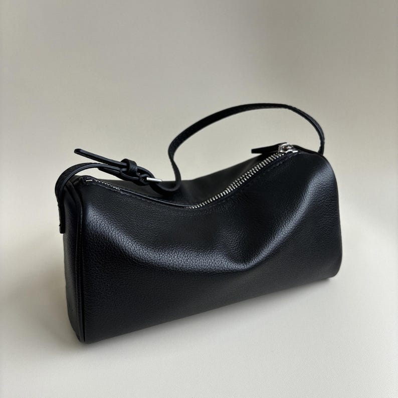 May include: A black, cylindrical handbag with a zipper closure and a small handle. The bag is made of a textured material and has a minimalist design. The zipper is silver-colored.