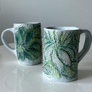 Puede incluir: Dos tazas de cerámica con base blanca y patrones florales en relieve de color verde y amarillo. Cada taza presenta un cuerpo cilíndrico, un asa curva y un acabado de superficie rugoso y artesanal con detalles de esmalte visibles.