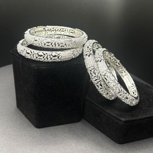 May include: Four silver bangles with a delicate floral design, set with sparkling white stones. The bangles are stacked on a black velvet display.