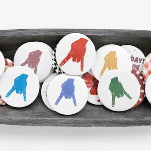 May include: A collection of white round buttons with a hand gesture design in various colors, including red, blue, and green. The buttons are arranged in a dark wooden bowl, with some patterned and text-printed buttons mixed in.