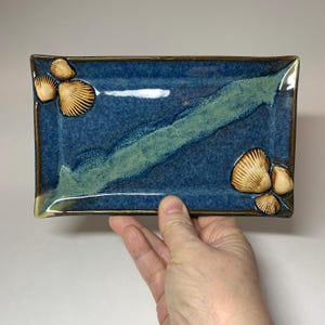 May include: A rectangular ceramic dish with a blue glaze and a green stripe across the center. The corners feature raised seashell designs in a tan color. The dish has a brown rim and is held by a hand.