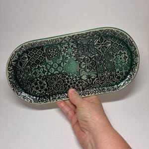 May include: An oval ceramic tray featuring a dark teal and black patchwork pattern of intricate geometric and floral motifs. The surface has a glossy finish with a raised edge, showcasing a variety of repeating tile-like designs in shades of green.