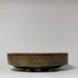 May include: A low, round ceramic bowl with a textured surface. The bowl has a dark brown rim and a green and brown body. It rests on three small, white feet. The bowl is likely for decorative or functional use.