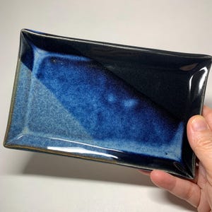 May include: A rectangular ceramic tray featuring a glossy glaze with a deep navy blue and black color gradient. The surface displays a smooth, reflective finish with subtle variations in tone, transitioning from a dark edge to a vibrant blue.