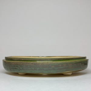 May include: A handcrafted ceramic bonsai pot. The oval-shaped pot features a textured, glazed surface with shades of green and brown. The interior is a light yellow color. The pot is elevated by three small, rounded feet.