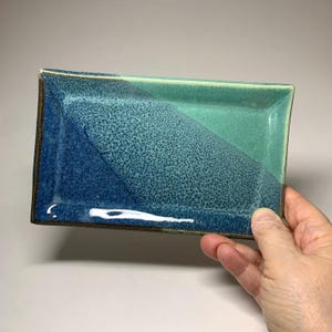 May include: A rectangular ceramic tray featuring a glossy glaze that transitions from a deep navy blue on one side to a vibrant turquoise green on the other. The surface displays a textured, speckled pattern where the two colors blend together.