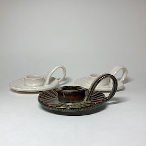 May include: Three ceramic candle holders. One is brown and green, with a handle and a ridged plate. The other two are white, with a handle and a ridged plate. The candle holders are on a white surface.