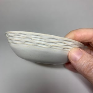 May include: A white ceramic bowl with a woven pattern around the rim. The bowl is slightly angled and has a speckled glaze.