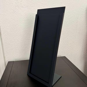 May include: A black, angled display stand with a rectangular frame and a small base. The stand is made of a matte black material and is designed to hold a sign or other display item. The stand is on a dark brown surface.